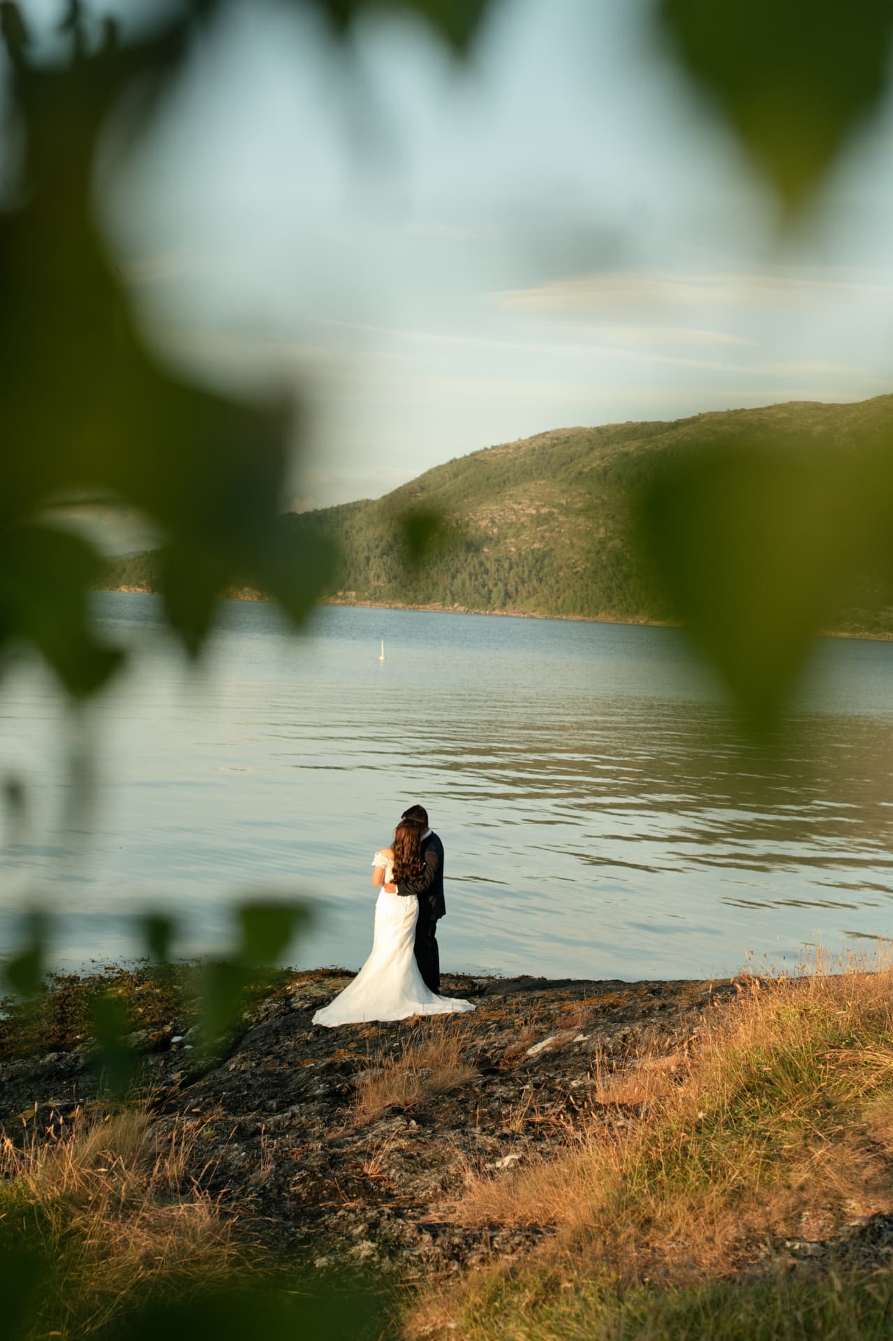 Couple embracing by the water, framed by green leaves